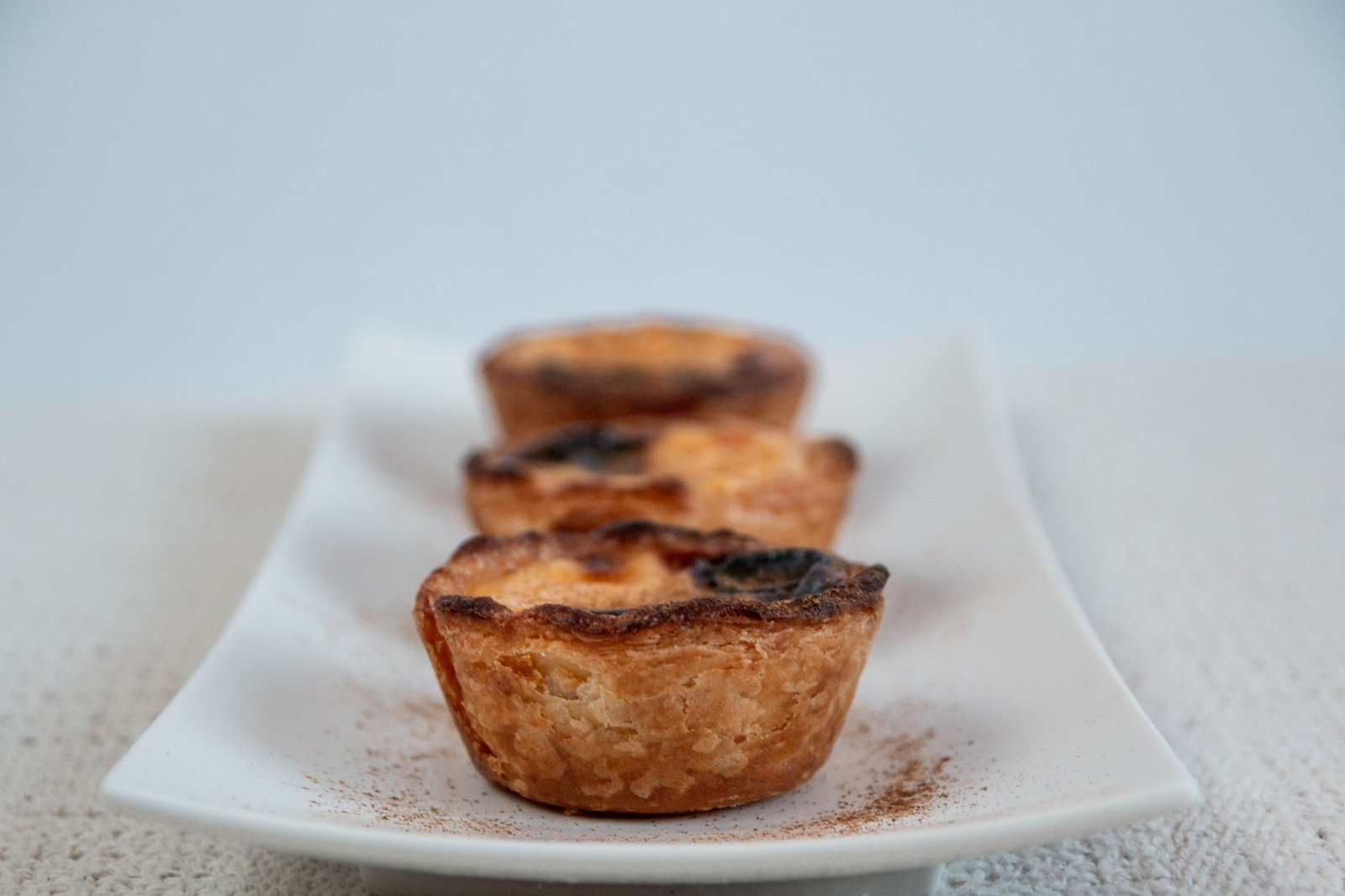 Trois pastéis de nata dorés et feuilletés sur une assiette blanche, saupoudrés de cannelle.
